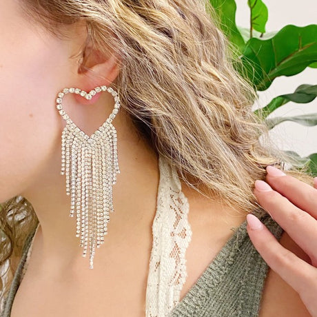 Heart-shaped earring with dangling beads worn by a person with greenery in the background