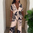 Woman wearing a beige and black floral pant set standing in front of a beige curtain with a plant to the side.